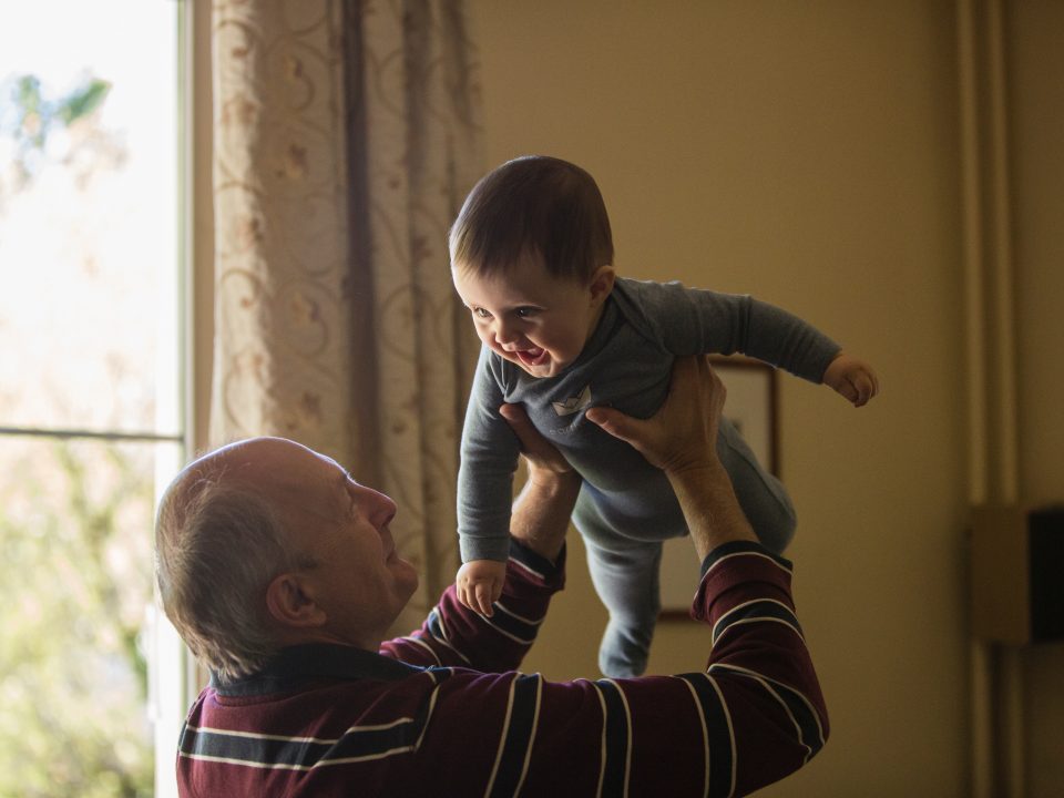 man wearing maroon, white, and blue stripe long-sleeved shirt lifting up baby wearing gray onesie