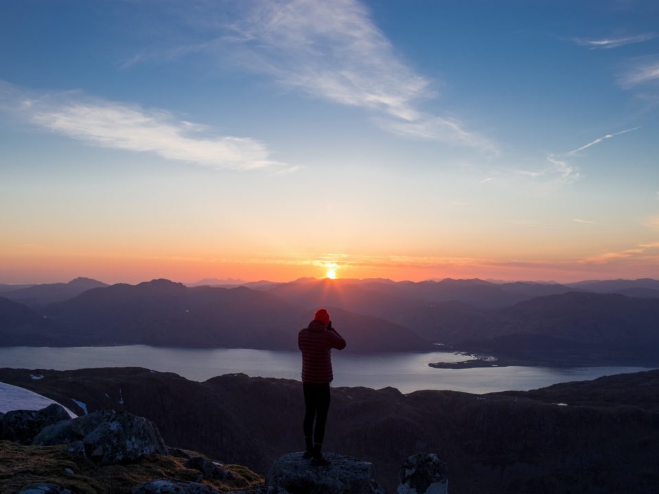 person standing on mountain ridge over lake under cloudy sky at sunset