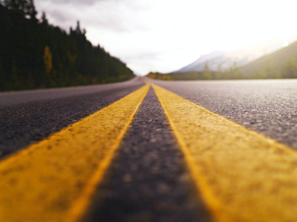 yellow and black road in between green trees under white clouds during daytime