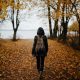 woman walking on pathway with falling leaves near body of water during daytime