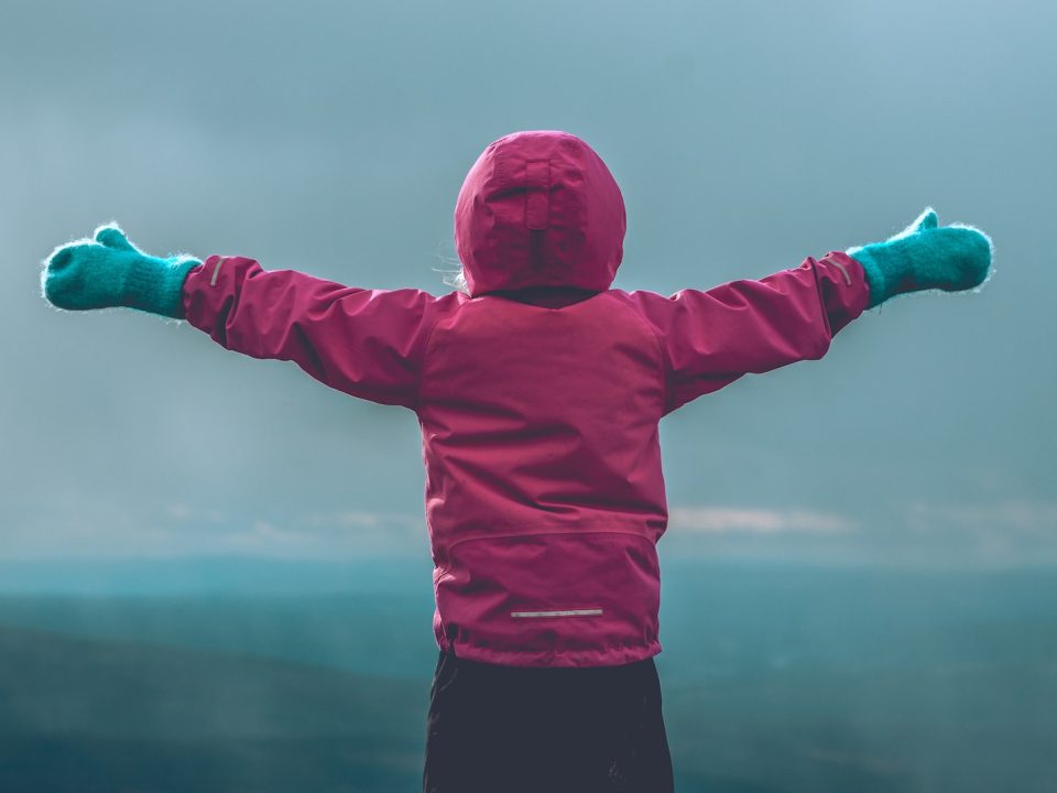 person wearing pink hooded jacket raising her hand in front of green mountain range during daytime