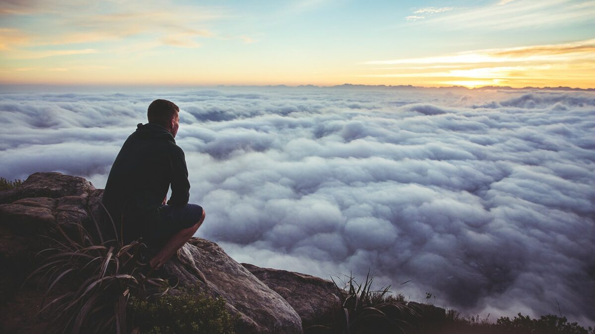 man sitting on gray rock while staring at white clouds