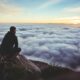 man sitting on gray rock while staring at white clouds