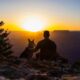 a man and his dog sitting on a cliff at sunset