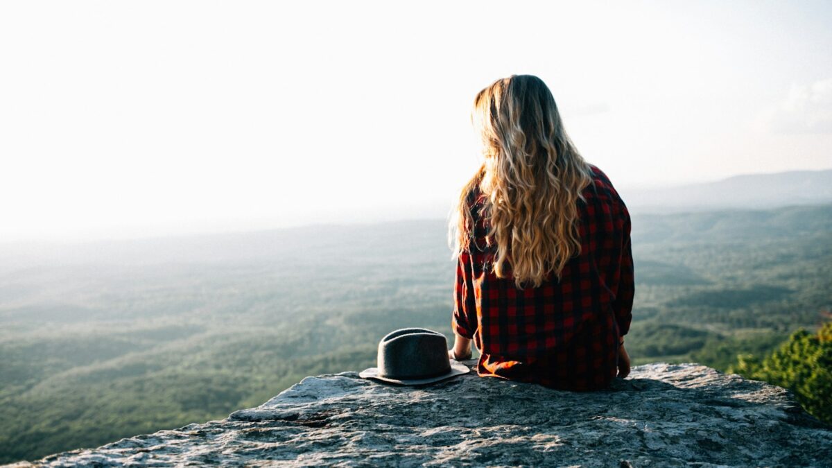 woman wearing red and black gingham shirt sitting on cliff with hat by its side