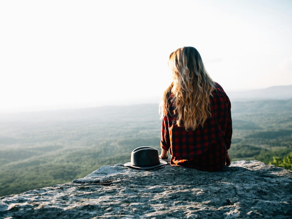 woman wearing red and black gingham shirt sitting on cliff with hat by its side