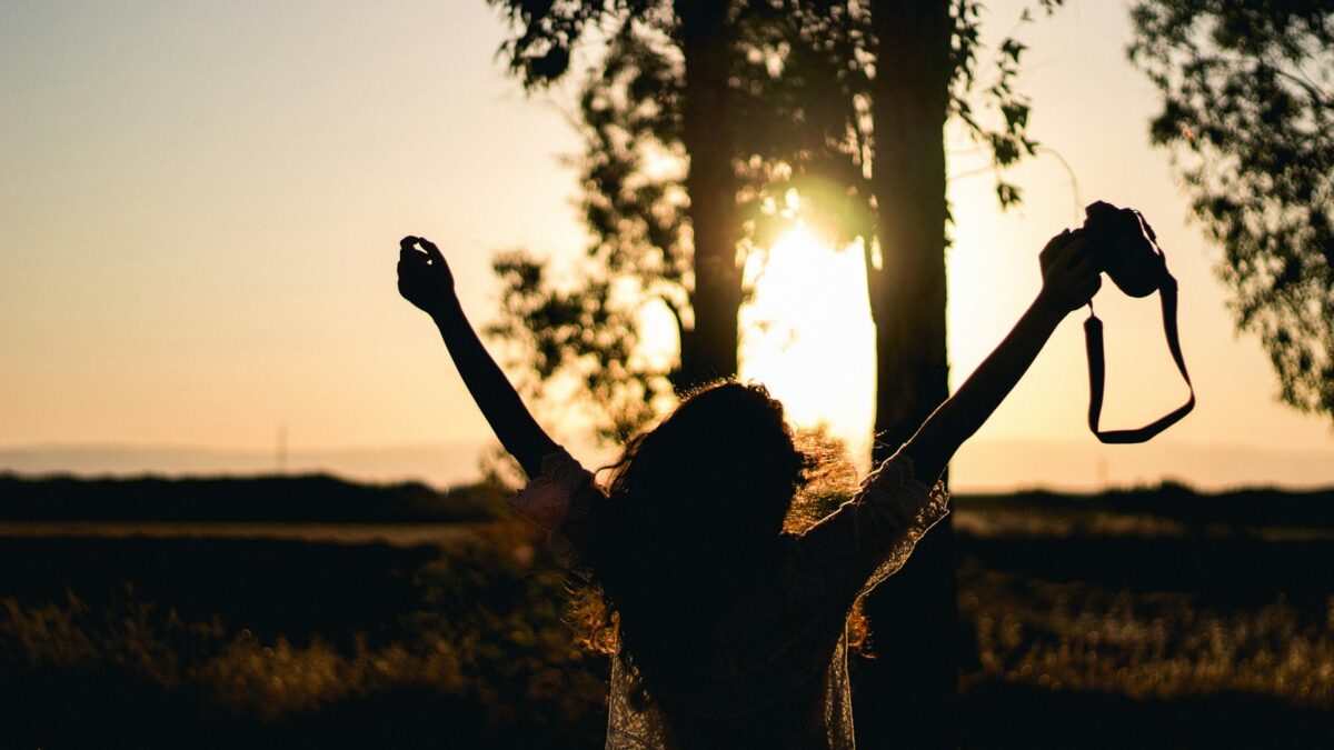 silhouette photo of woman raising her hands while holding DSLR camera