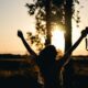 silhouette photo of woman raising her hands while holding DSLR camera