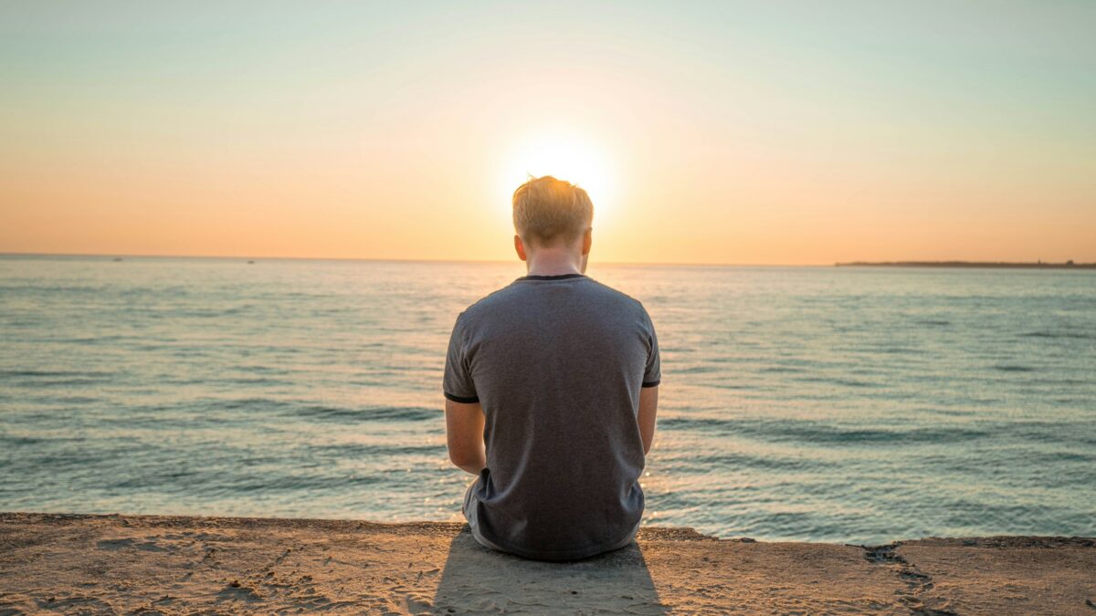 man sitting on sand front of sea during golden hour