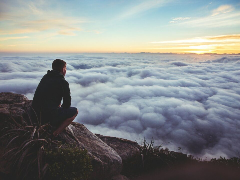 man sitting on gray rock while staring at white clouds